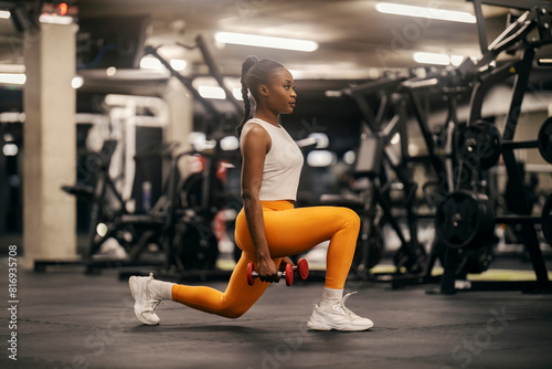 Side view of a strong african american sportswoman doing lunges with dumbbells at gym.