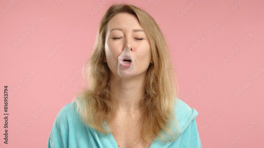 Captured in a lighthearted moment, a young woman in a sky-blue robe chews bubble gum and blows a big bubble against a pastel pink backdrop. 