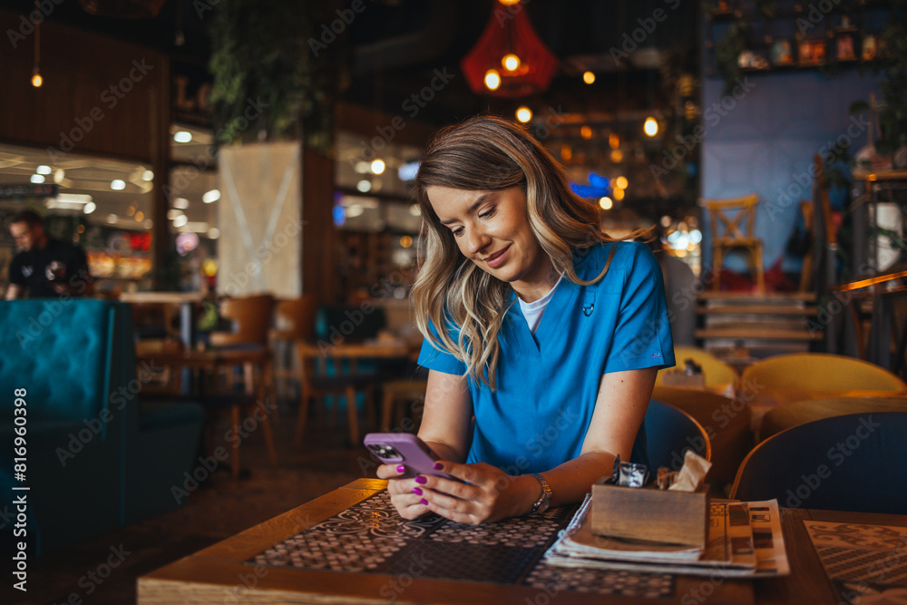 A female nurse in blue scrubs enjoys a relaxing moment, browsing her phone at a cozy cafe table surrounded by warm ambient lighting. A female nurse in blue scrubs enjoys a relaxing moment, browsing her phone at a cozy cafe table surrounded by warm ambient lighting.