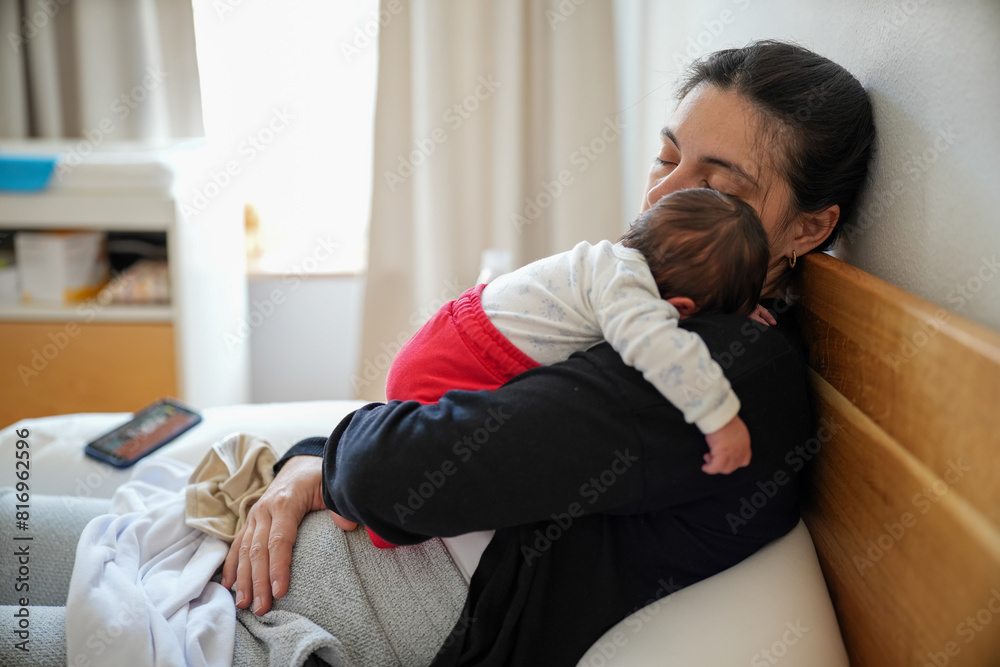 Mother resting on a bed with her newborn baby sleeping on her chest. The baby, dressed in a white top and red pants, highlights the peaceful and tender bond between them. Mother resting on a bed with her newborn baby sleeping on her chest. The baby, dressed in a white top and red pants, highlights the peaceful and tender bond between them.