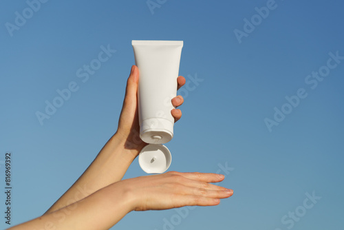 Female hands apply cream from white jar mockup on hand against blue sky background. Concept of skin care, nutrition and hydration, beauty and spa.