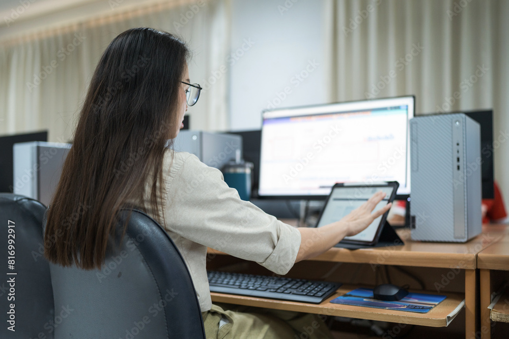 A woman sits at a desk in front of a computer. She is wearing glasses and is focused on her work. The room is filled with other people working on their computers, creating a busy