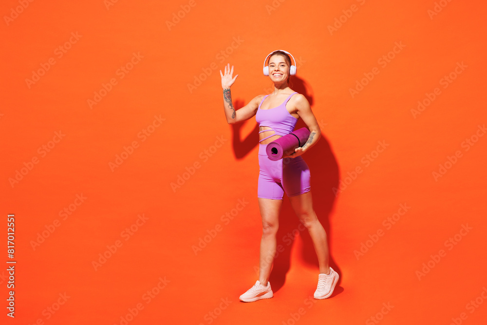 Full body young fitness trainer woman sportsman in top shorts purple clothes headphones train in home gym hold yoga mat listen music isolated on plain orange background Workout sport fit abs concept