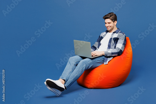 Full body happy young IT man he wear shirt white t-shirt casual clothes sit in bag chair hold use work on laptop pc computer isolated on plain blue cyan background studio portrait. Lifestyle concept.