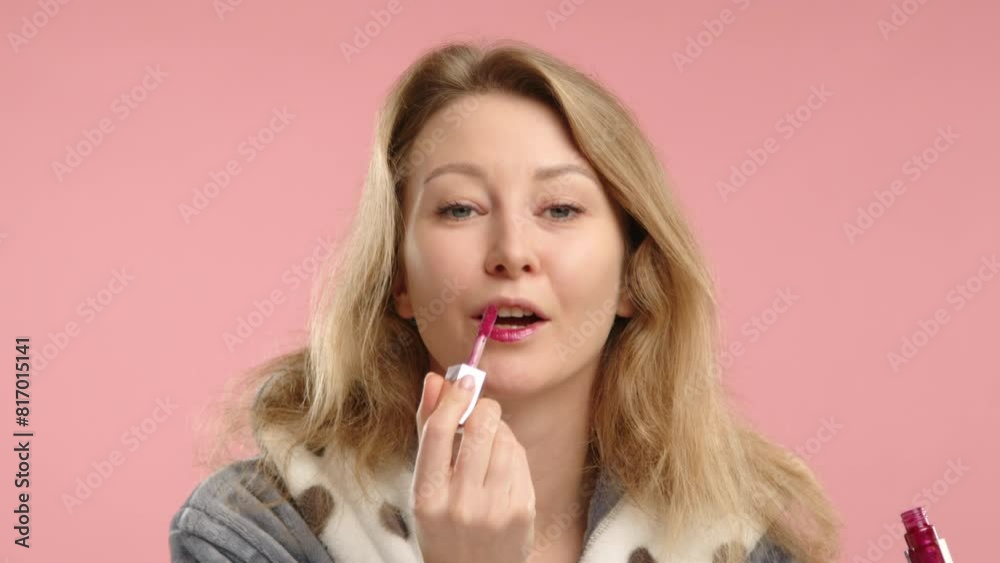 An attractive young woman in a cozy robe applies pink lipstick with precision, set against a minimalist pink background that highlights her beauty routine and personal grooming habits. An attractive young woman in a cozy robe applies pink lipstick with precision, set against a minimalist pink background that highlights her beauty routine and personal grooming habits.