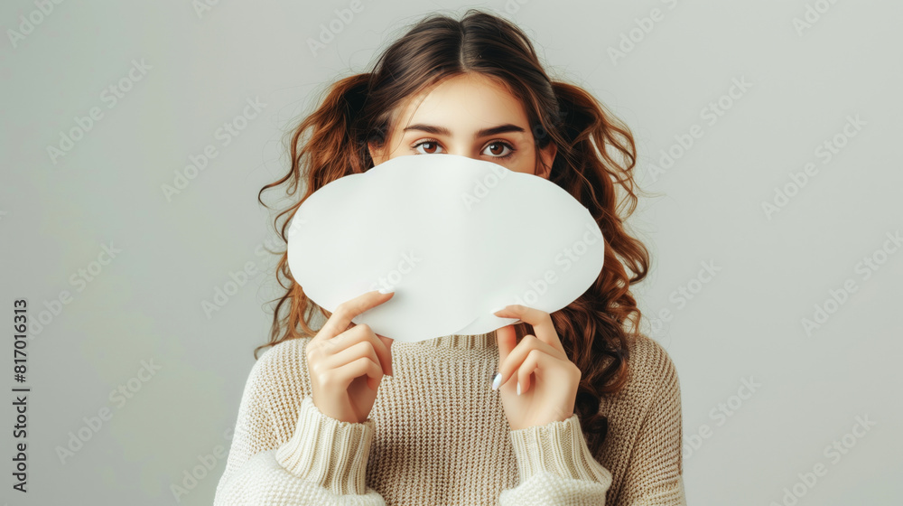 Portrait of a young woman with curly hair holding a blank white paper in front of her face, with only her eyes visible. She is wearing a cozy knitted sweater. Portrait of a young woman with curly hair holding a blank white paper in front of her face, with only her eyes visible. She is wearing a cozy knitted sweater.