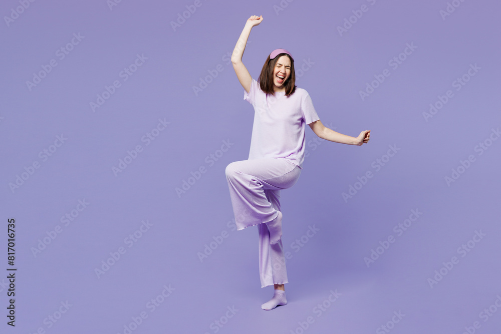Full body young woman wear pyjamas jam sleep eye mask rest relax at home hold pillow do winner gesture clench fists raise up leg dance isolated on plain purple background. Good mood night nap concept.