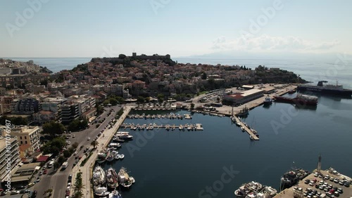 Glide above the stunning city of Kavala in Greece, witnessing boats smoothly docked in the harbor against a backdrop of architectural beauty and shimmering waters below