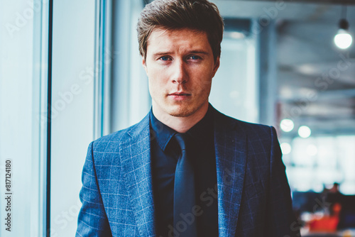 Half length portrait of serious expert economist in elegant suit keeping hands in pockets, stylish confident businessman in formal wear looking at camera while standing in modern company office photo