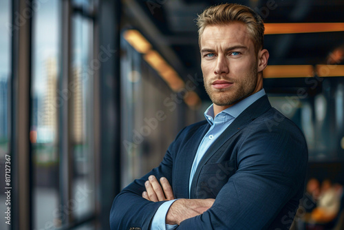In a sleek office, a blond, blue-eyed man, muscular and dapper in a suit, stands confidently