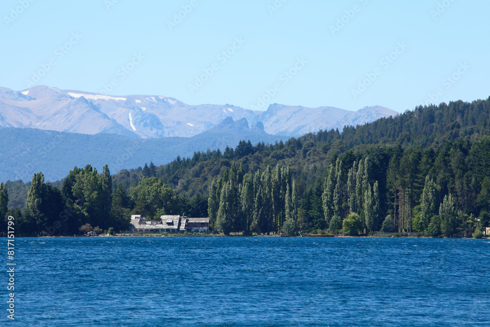 dense green forest on a mountainside near a blue lake with an old building on the coast and mountain peaks in the distance