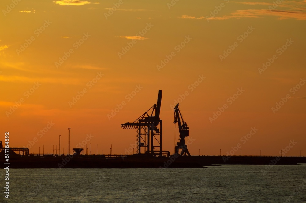 Obraz premium Silhouetted Industrial Cranes at Sunset, Creating a Dramatic and Peaceful Scene at the End of the Workday, Capturing the Beauty of Industrial Landscapes.