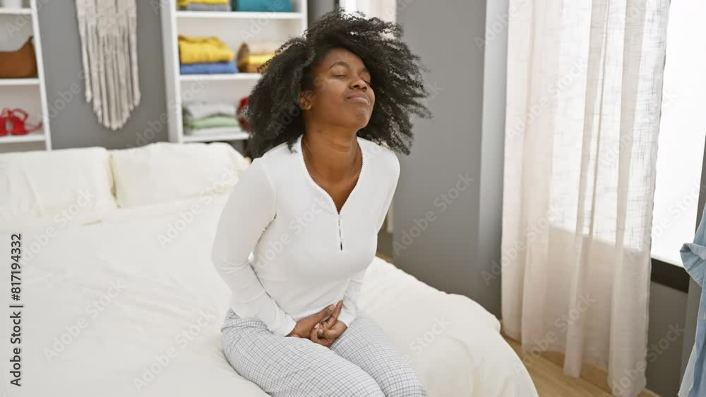 An african american woman in pajamas feeling stomach pain while sitting on a bed in a home setting An african american woman in pajamas feeling stomach pain while sitting on a bed in a home setting