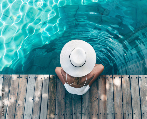 Top view of a woman sitting by a clear blue pool, wearing a white swimsuit and hat at the poolside