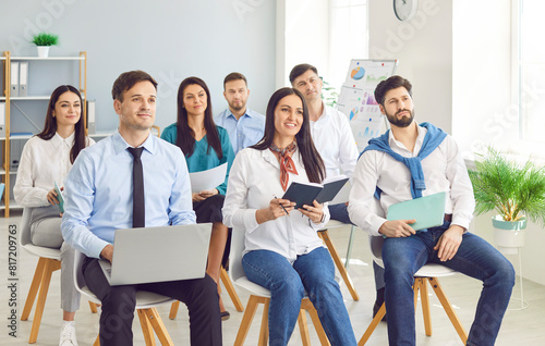 Group of smiling young business people sitting in a row on chairs and listening to colleague or their leadership at working place during business training or conference in meeting room.