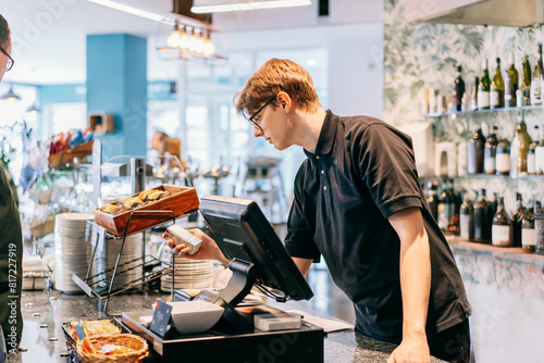 Young waiter serving customer at cash point in cafe. Man working with POS terminal. Cashier, barista checking for payment receipt. Hospitality, server and preparing a slip at the till in coffee shop. photo