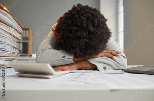 Tired burned out African American woman hides her face in hands and lays weary head with Afro hair on desk, exhausted by red tape paperwork piles, feeling stuck in this goddamn office. Burnout concept photo