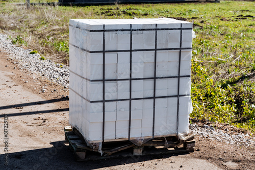Stacked bundle of white construction bricks on a wooden pallet outdoors, secured with plastic wrap and black straps. Ready for building project at a nonresidential construction site photo