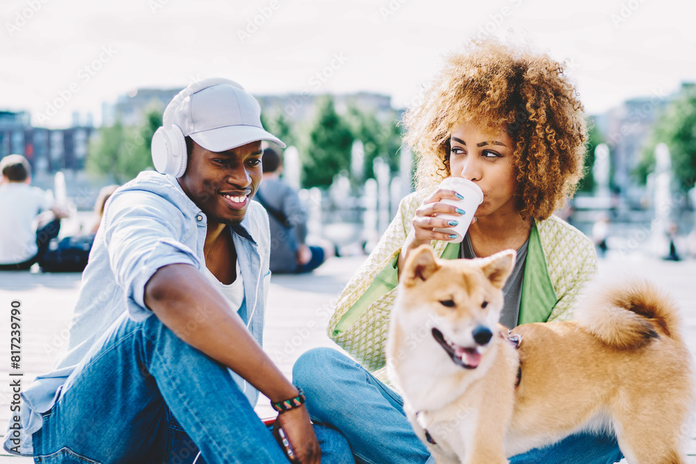 Positive dark skinned couple in love dressed in casual wear having fun and spending free time together with dog in urban setting.Cheerful african american young man and woman drinking coffee to go