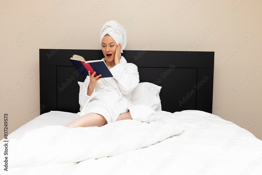 Adult Woman Sits In Bed with Surprised, Shocked Facial Expression while Reading A Book. Emotional Expression Of Face, Caucasian Mature Female Reading Horror Novel, Fiction, Detective Story. Book Lover Adult Woman Sits In Bed with Surprised, Shocked Facial Expression while Reading A Book. Emotional Expression Of Face, Caucasian Mature Female Reading Horror Novel, Fiction, Detective Story. Book Lover