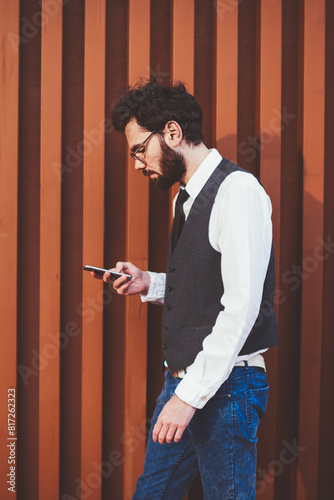 Stylish man walking on city street with telephone in hand and reading sms message from friend, caucasian hipster guy using 4g connection for searching information on modern smartphone device