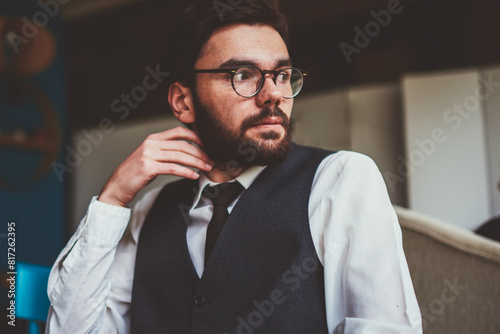 Thoughtful hipster guy in spectacles for vision correction sitting at cafeteria table with coffee cup and pondering on leisure, young pensive man in trendy apparel waiting friend at cafe interior