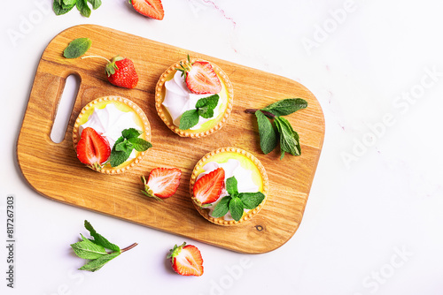Fresh homemade tartlets with lemon decorated with aromatic strawberries and green mint leaves on the serving board on white marble table top view.