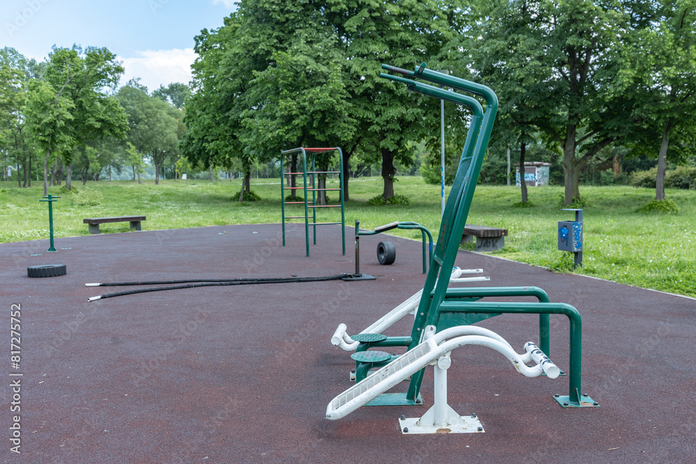 Obraz premium An outdoor fitness park featuring a variety of exercise equipment such as pull-up bars, leg press machine, and push-up bars, set on a rubber flooring surface with a backdrop of trees.