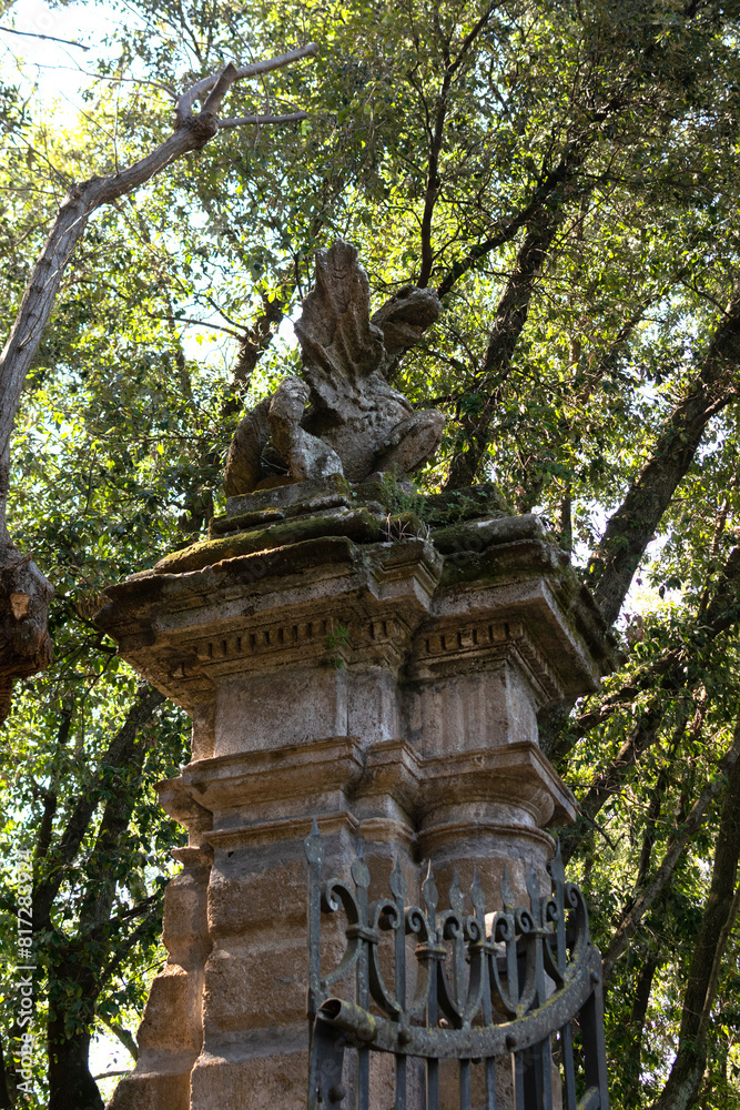 In the summer park there is an ancient stone high gate with an iron lattice, on top there is a statue of a dragon, vertical view
