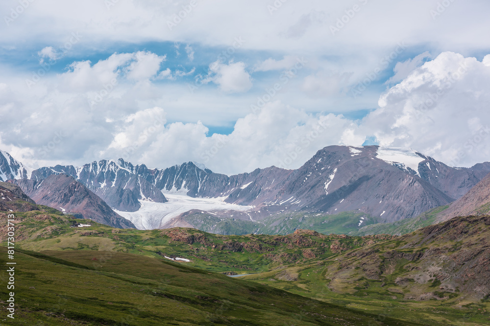 Scenic alpine landscape with green hills and rocks with view to big glacier and large snow-capped mountain top far away under clouds in blue sky in changeable weather. Awesome high snowy mountains. Scenic alpine landscape with green hills and rocks with view to big glacier and large snow-capped mountain top far away under clouds in blue sky in changeable weather. Awesome high snowy mountains.