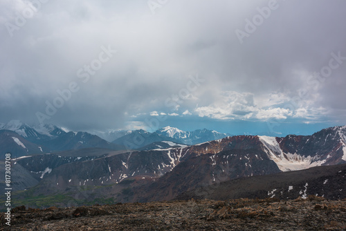 Dramatic alpine top view to rocky ridge and large mountain range with most beautiful snow-capped peak in sunlight in gray rainy low clouds. Scenic landscape with big snowy top in rain grey cloudy sky.