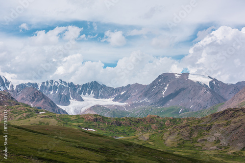 Scenic alpine landscape with green hills and rocks with view to big glacier and large snow-capped mountain top far away under clouds in blue sky in changeable weather. Awesome high snowy mountains.