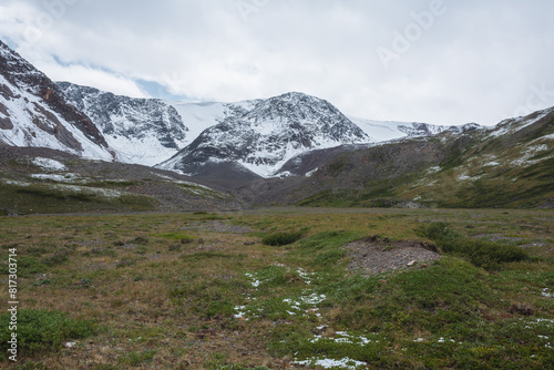Dramatic cold landscape with green valley against snow covered large mountain range under rainy cloudy gray sky. Gloomy rainy weather in high mountains. Alpine view to big snowy mount in rain time.