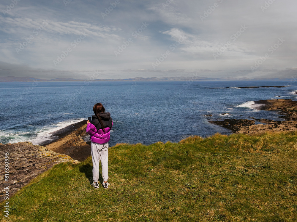 A girl in a purple jacket stands on grass . She is wearing gray sweatpants and. The ocean is in the background and the sky is cloudy and blue. Travel and tourism. Mullaghmore, Ireland.