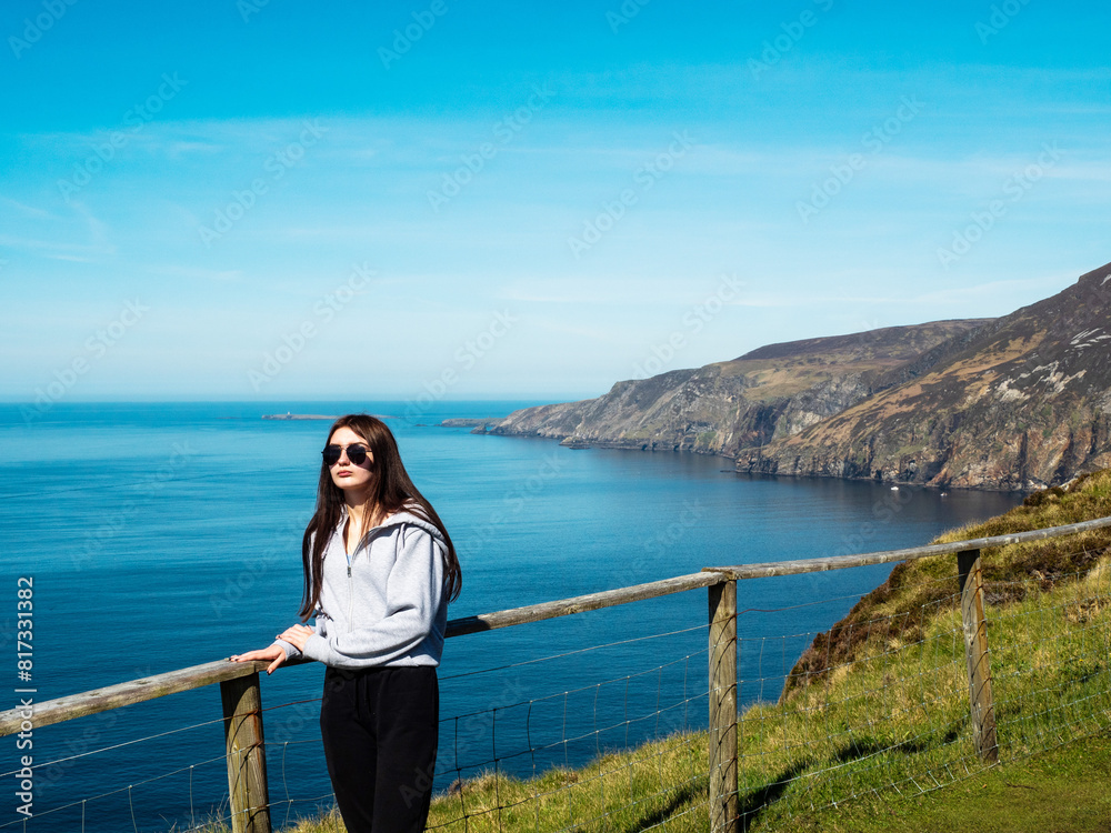 Teenager girl on a trip to Slieve League Cliff, Ireland. Stunning nature scenery with cliff, ocean and clean blue sky. Travel and tourism concept. Model posing in nature scene. Sunny day. Teenager girl on a trip to Slieve League Cliff, Ireland. Stunning nature scenery with cliff, ocean and clean blue sky. Travel and tourism concept. Model posing in nature scene. Sunny day.
