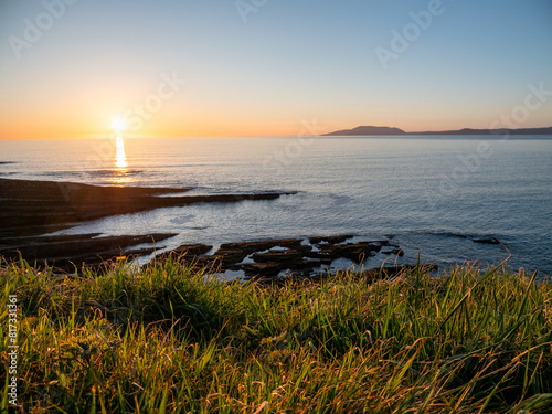 Stunning sunset nature scene with rough stone coastline, green grass hill, dramatic sky and mountains in the background. Mullaghmore head area, Ireland. Sun flare. Irish landscape.