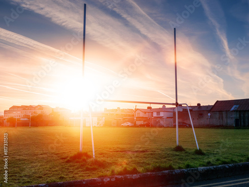 Tall goal post in a field at sun rise. Cloudy sky with sun flare. Irish sport training ground in a city. Nobody. Hurling, rugby and camogie practice area and national sport in Ireland.