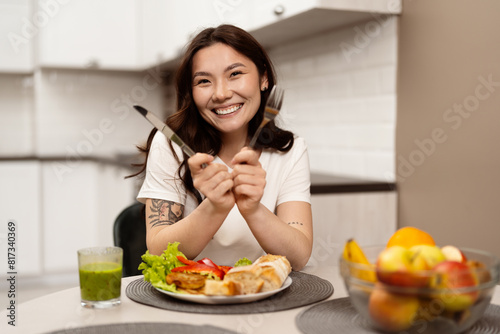 Happy Young Woman Enjoying A Healthy Meal In A Modern Kitchen, Smiling Joyfully With Fork And Knife, Fresh Food On Table.