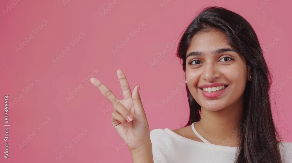 Portrait of beautiful young South Asian woman wearing white top smiling and showing the peace sign on a pink background, template for advertising or promotion, with copy space for text Portrait of beautiful young South Asian woman wearing white top smiling and showing the peace sign on a pink background, template for advertising or promotion, with copy space for text