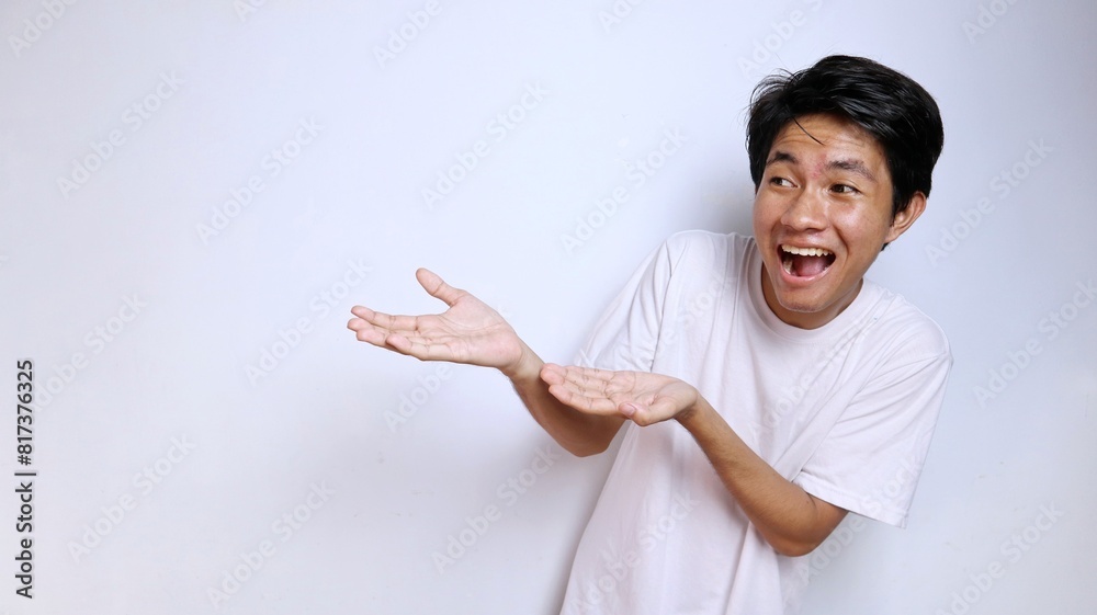 Young Asian man in white shirt with funny expression gestures with open palm towards copy space Young Asian man in white shirt with funny expression gestures with open palm towards copy space