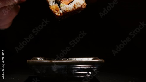 Hand using chopsticks dipping sushi roll into soy sauce in gravy boat on black background close-up. photo