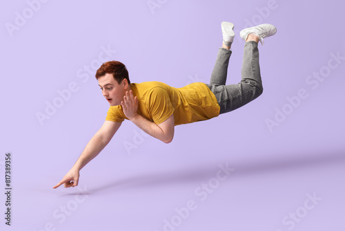 Obraz na plátně Surprised young man flying and pointing at something on lilac background