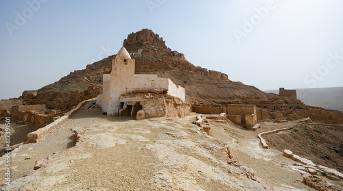 Сrumbling ruins of ancient deserted village of Ksar Guermessa standing atop hills in Tataouine with preserved white mosque in golden arid Tunisian landscape photo