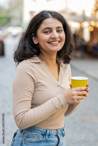 Smiling happy Indian young woman enjoying morning coffee hot drink outdoors. Relaxing taking a break. Girl drinking coffee to go walking passes the urban city street. Town lifestyles outside. Vertical