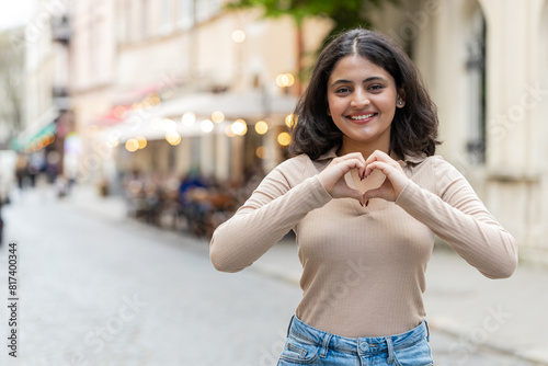 I love you. Indian young woman makes symbol of love, showing heart sign to camera, express romantic feelings, express sincere positive feelings. Charity, gratitude, donation. Girl on urban city street