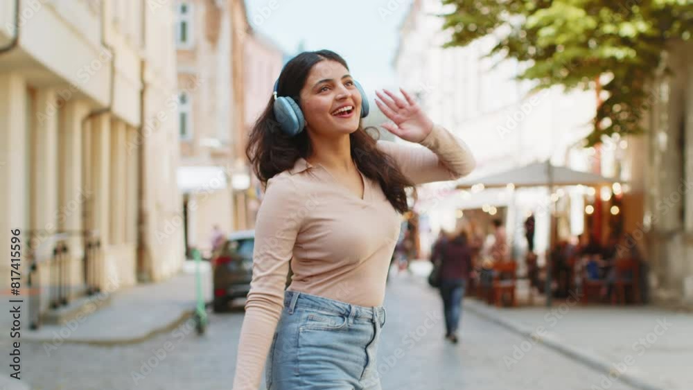 Back view of happy Indian young woman in wireless headphones dancing listening favorite energetic disco music in smartphone dancing outdoors. Hispanic girl walking on urban city street. Town lifestyle Back view of happy Indian young woman in wireless headphones dancing listening favorite energetic disco music in smartphone dancing outdoors. Hispanic girl walking on urban city street. Town lifestyle