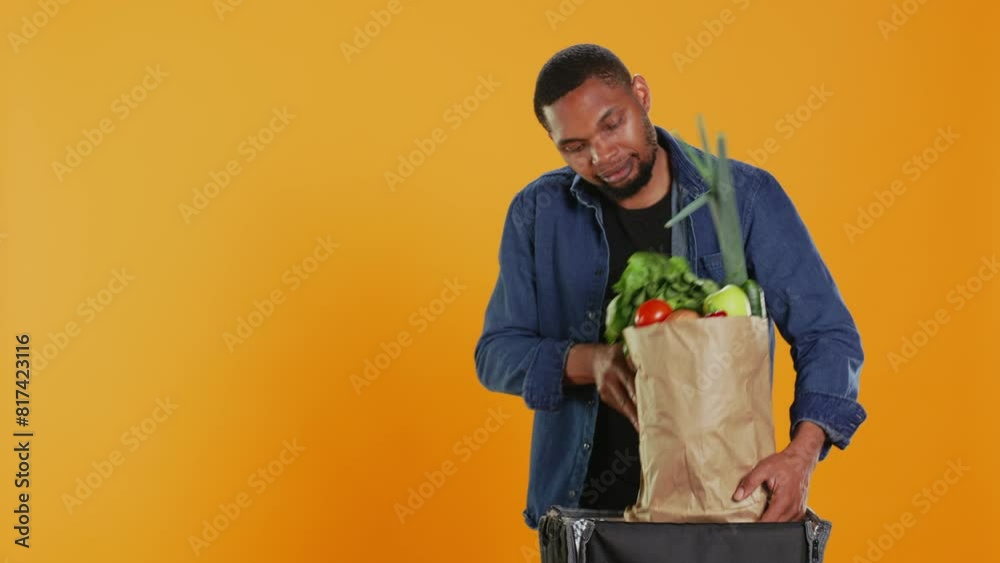 Young takeout employee delivering a paper bag full of bio produce in a thermal backpack, bringing local supermarket order to customers. Male courier carrying organic fruits and vegetables. Camera B. Young takeout employee delivering a paper bag full of bio produce in a thermal backpack, bringing local supermarket order to customers. Male courier carrying organic fruits and vegetables. Camera B.