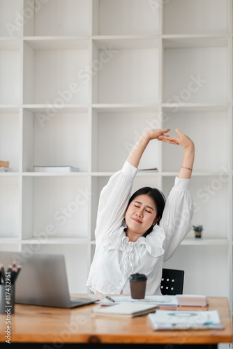A woman is stretching her arms and legs while sitting at a desk with a laptop and a cup