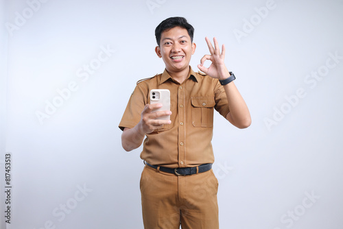 Male Asian civil servant in brown uniform holding a smartphone and showing okay sign photo