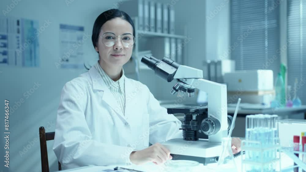 Young female scientist in medical coat sitting at desk in laboratory, using microscope and posing at camera. Video portrait